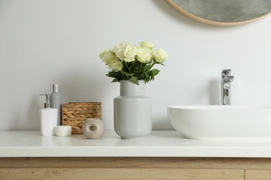 Vase with beautiful white roses and toiletries near sink in bathroom