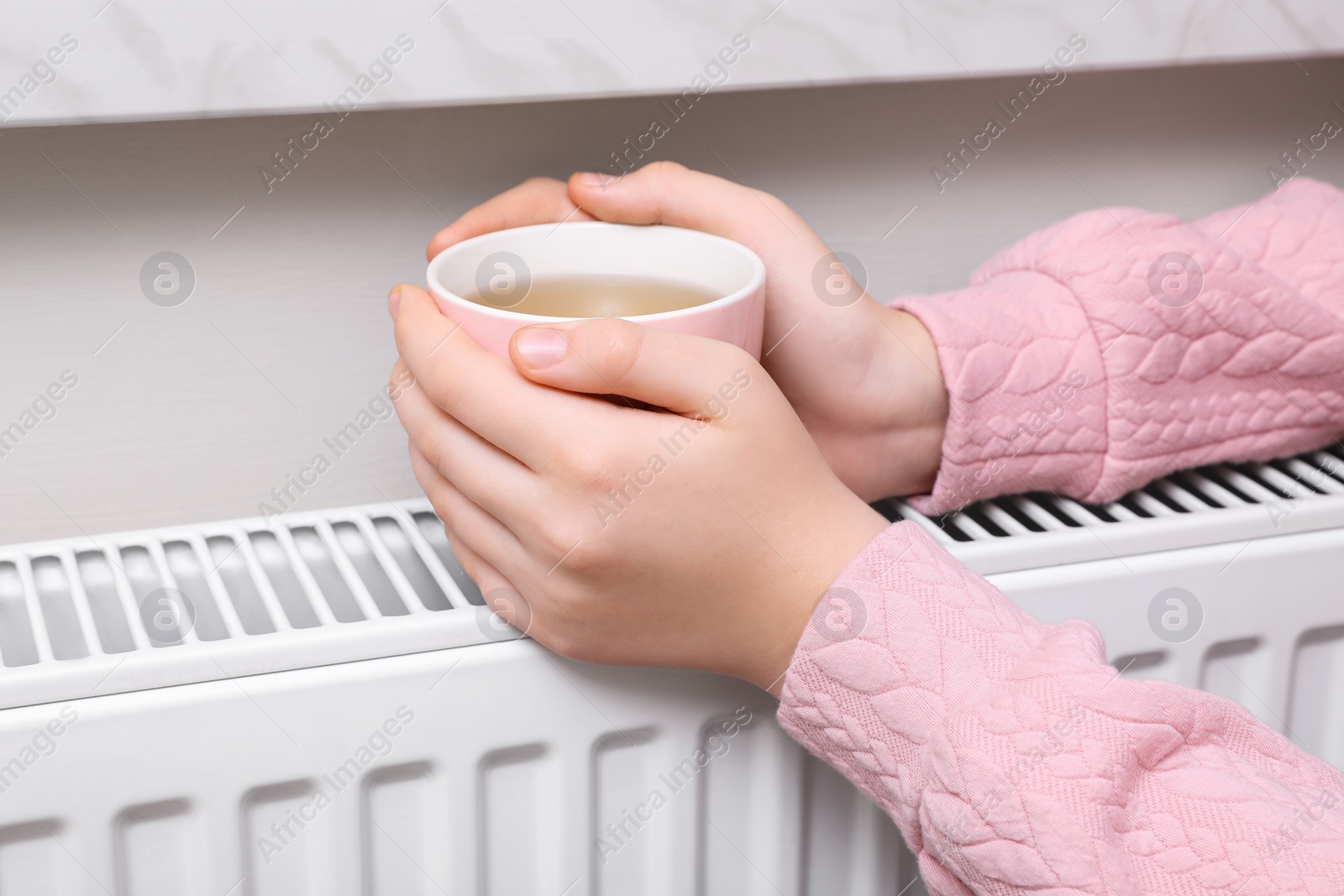 Photo of Girl with cup of tea warming hands on heating radiator indoors, closeup