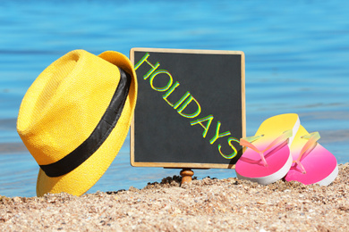 Image of Little blackboard with word HOLIDAYS, hat and flip flops on sand near sea