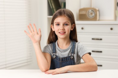 Happy little girl waving hello at white table indoors