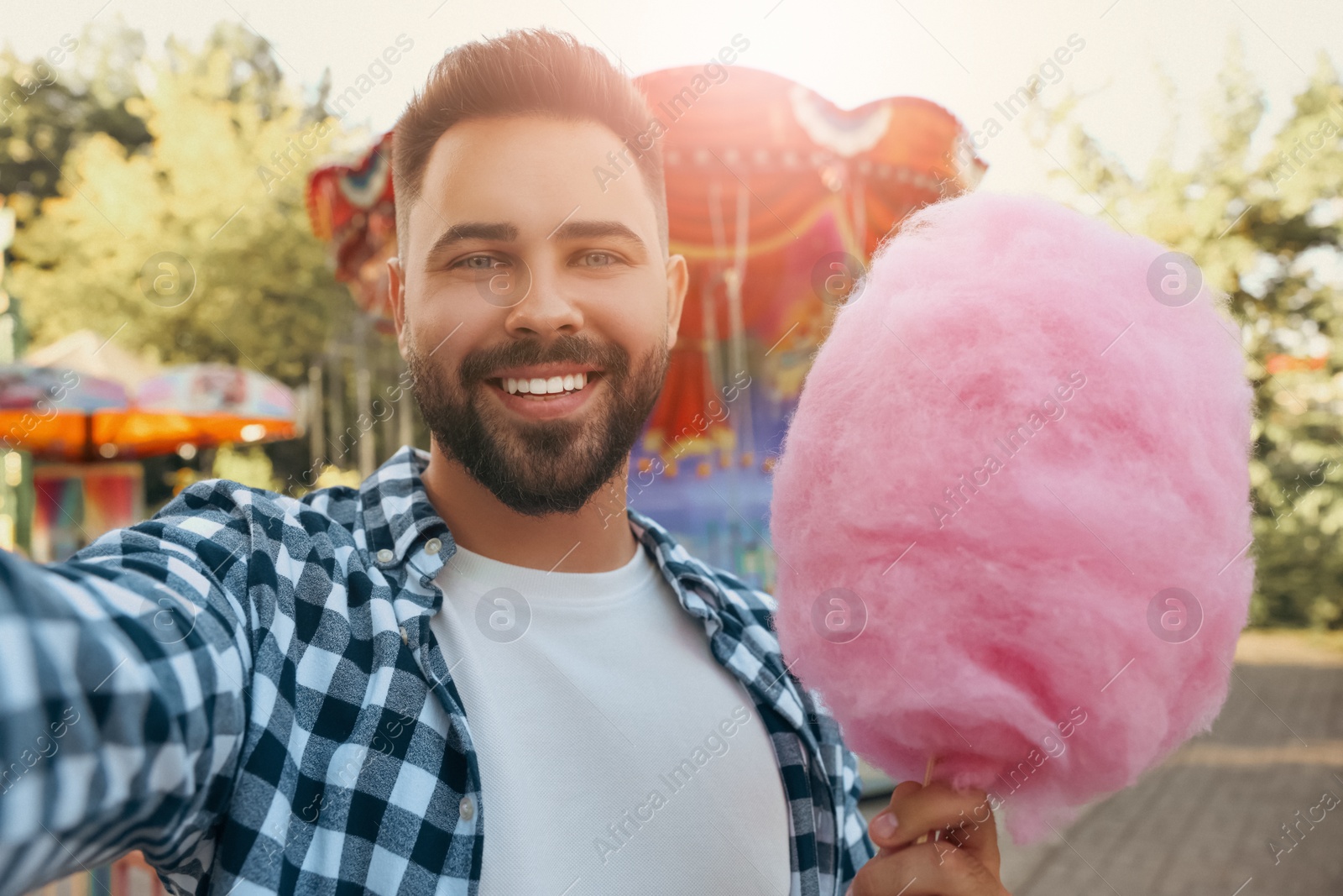 Image of Happy young man with cotton candy taking selfie at funfair on sunny day