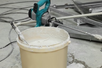 Photo of Bucket with plaster and putty knife near construction equipment on concrete floor