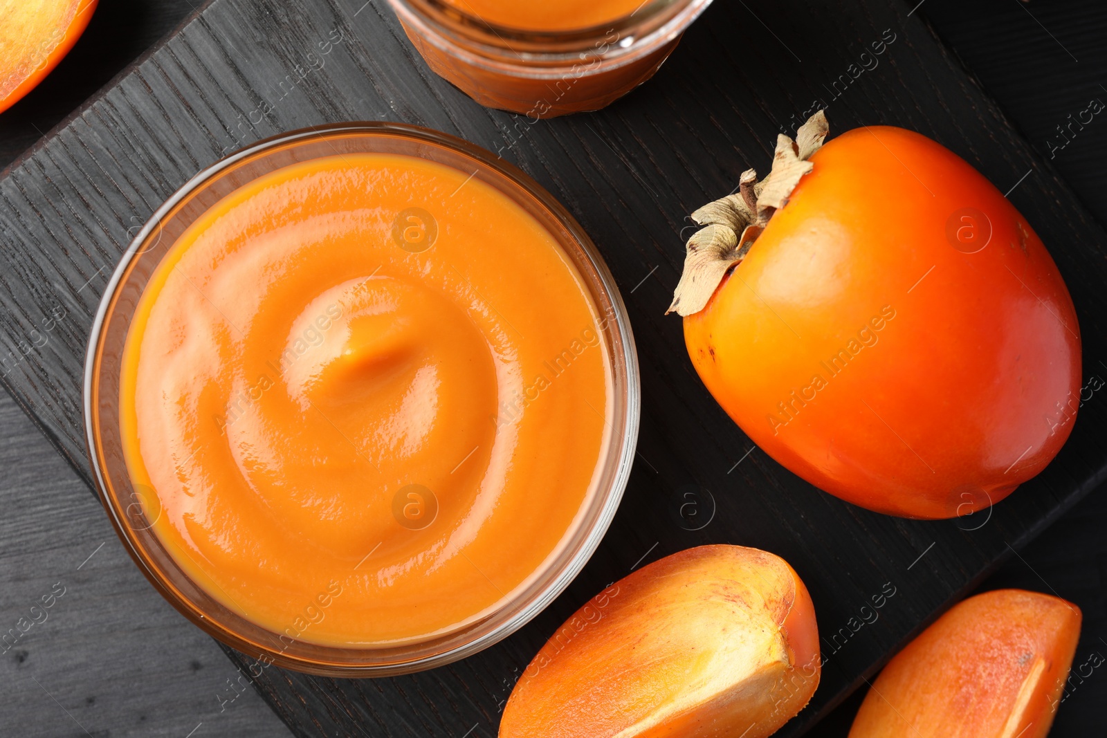 Photo of Delicious persimmon jam in glass bowl and fresh fruits on dark gray wooden table, top view