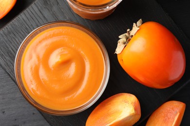 Photo of Delicious persimmon jam in glass bowl and fresh fruits on dark gray wooden table, top view