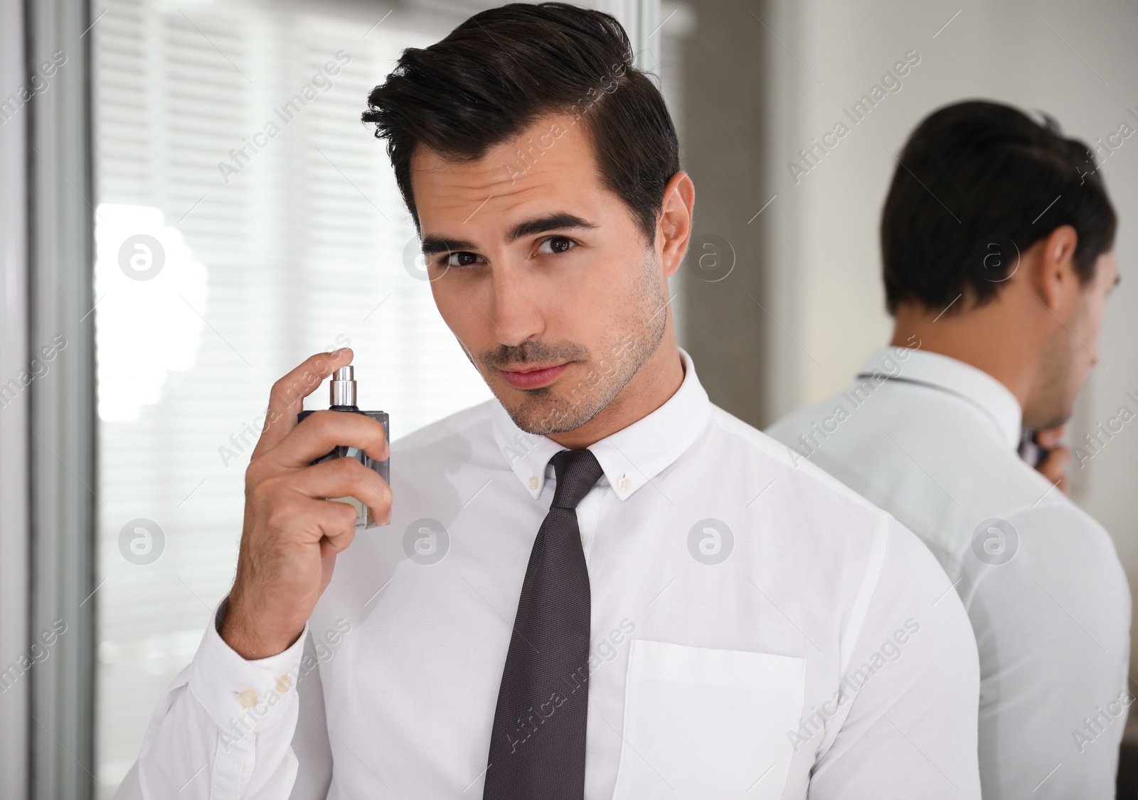 Photo of Handsome young man using perfume near mirror indoors
