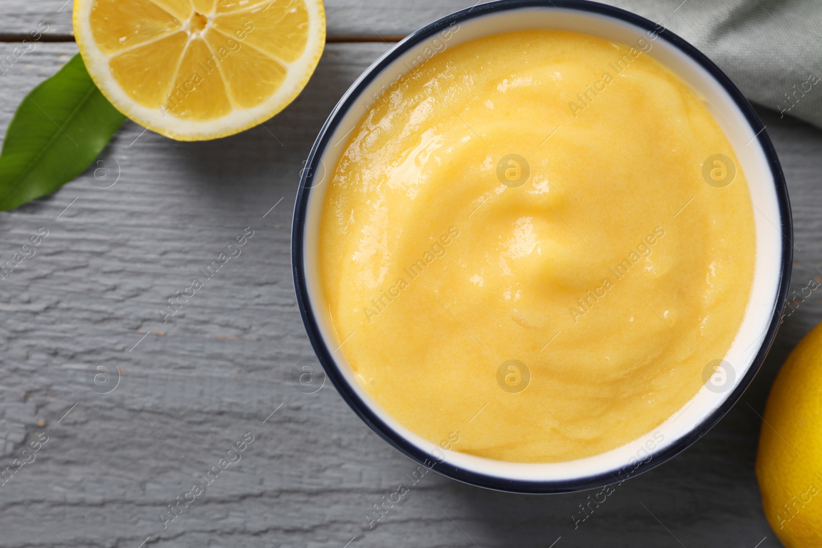 Photo of Delicious lemon curd in bowl, fresh citrus fruit and green leaf on grey wooden table, top view. Space for text