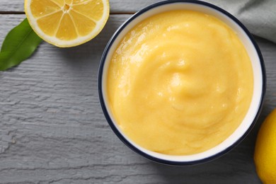 Photo of Delicious lemon curd in bowl, fresh citrus fruit and green leaf on grey wooden table, top view. Space for text