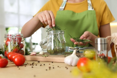 Woman putting herbs into pickling jar at table in kitchen, closeup