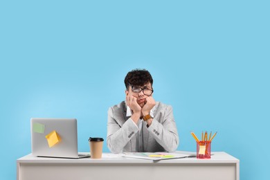 Photo of Tired young man working at white table on light blue background. Deadline concept
