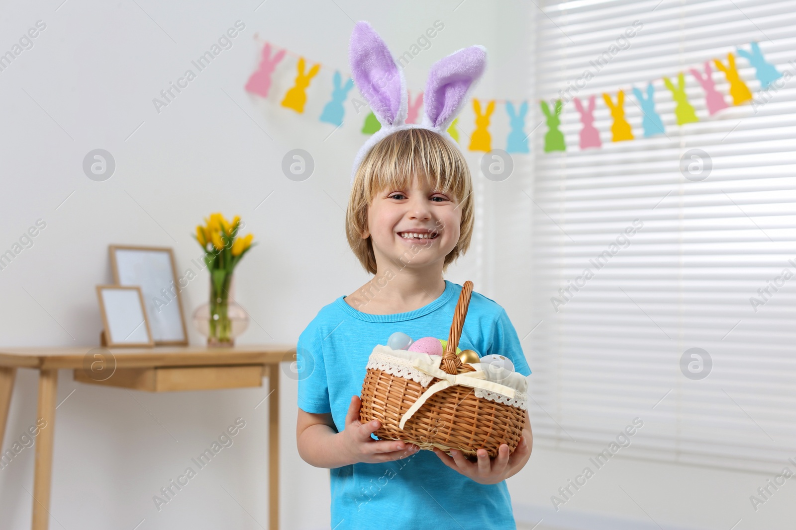 Photo of Happy boy in bunny ears headband holding wicker basket with painted Easter eggs indoors