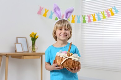 Happy boy in bunny ears headband holding wicker basket with painted Easter eggs indoors