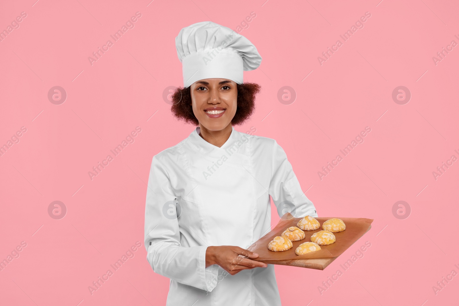 Photo of Happy female chef in uniform holding board with cookies on pink background