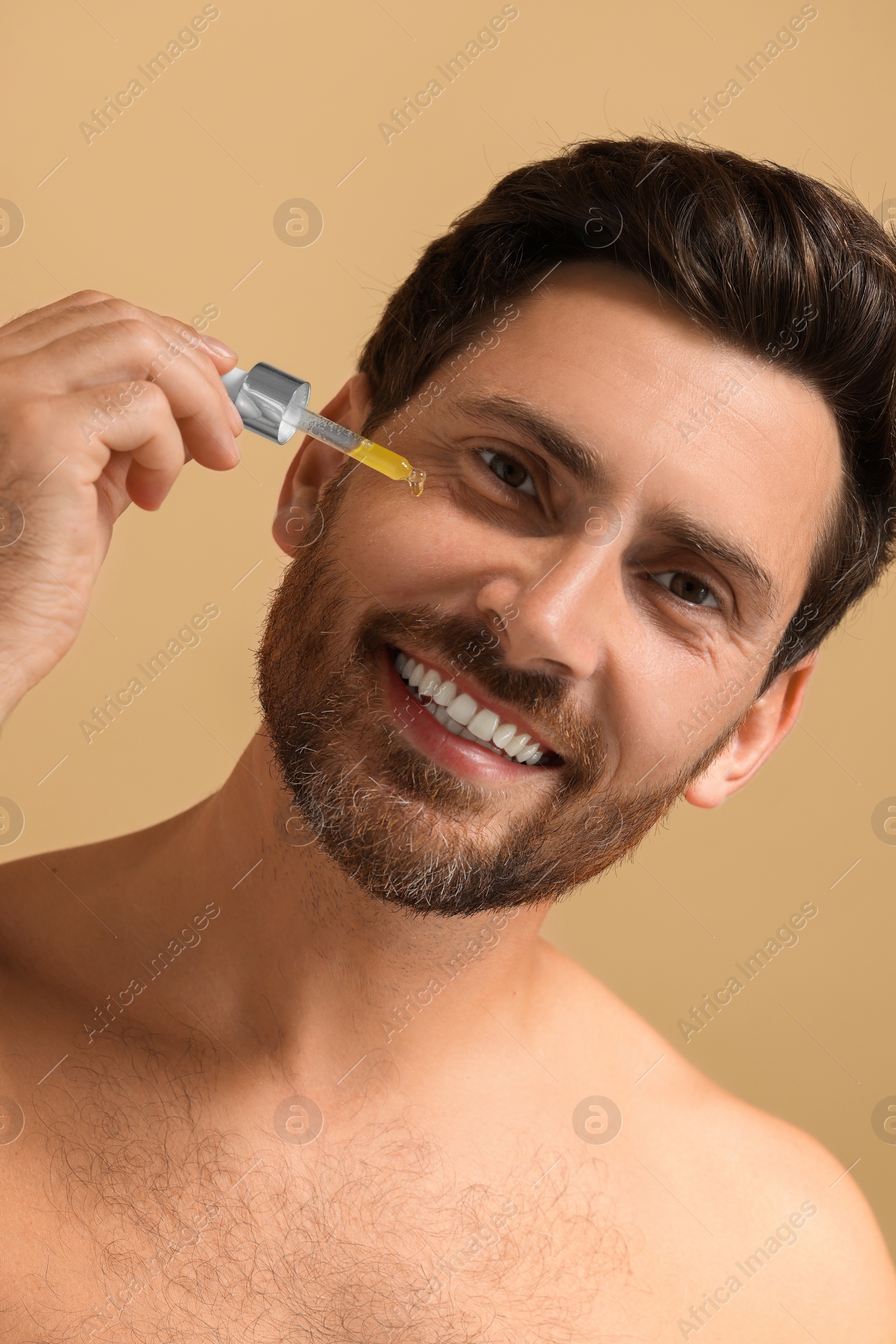 Photo of Smiling man applying cosmetic serum onto his face on beige background, closeup