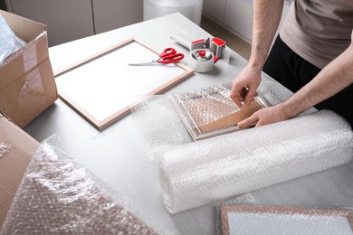 Man covering photo frame with bubble wrap at light grey table, closeup