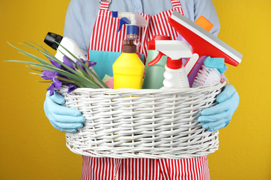 Photo of Woman holding basket with spring flowers and cleaning supplies on yellow background, closeup