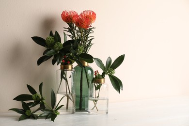 Photo of Vases with beautiful flowers and branches on white wooden table near beige wall. Space for text