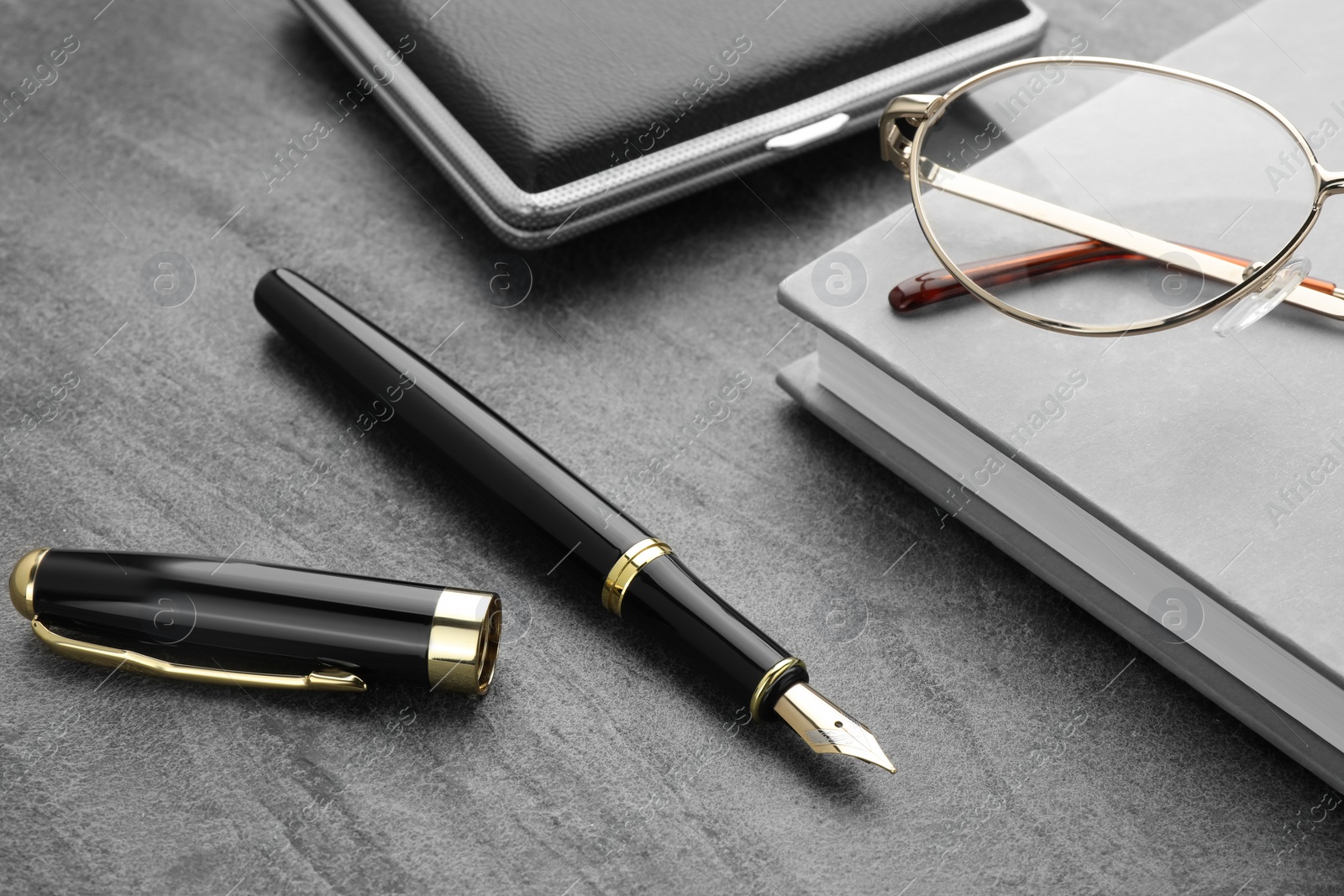 Photo of Stylish black fountain pen, glasses, notebook and cigarette case on grey textured table, closeup