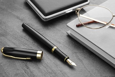 Stylish black fountain pen, glasses, notebook and cigarette case on grey textured table, closeup