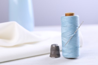 Photo of Spool of light blue sewing thread with needle and thimble on white table, space for text