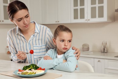 Photo of Mother feeding her daughter in kitchen. Little girl refusing to eat dinner