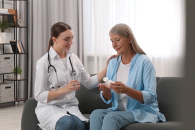 Young healthcare worker giving glass of water to senior woman with pills indoors