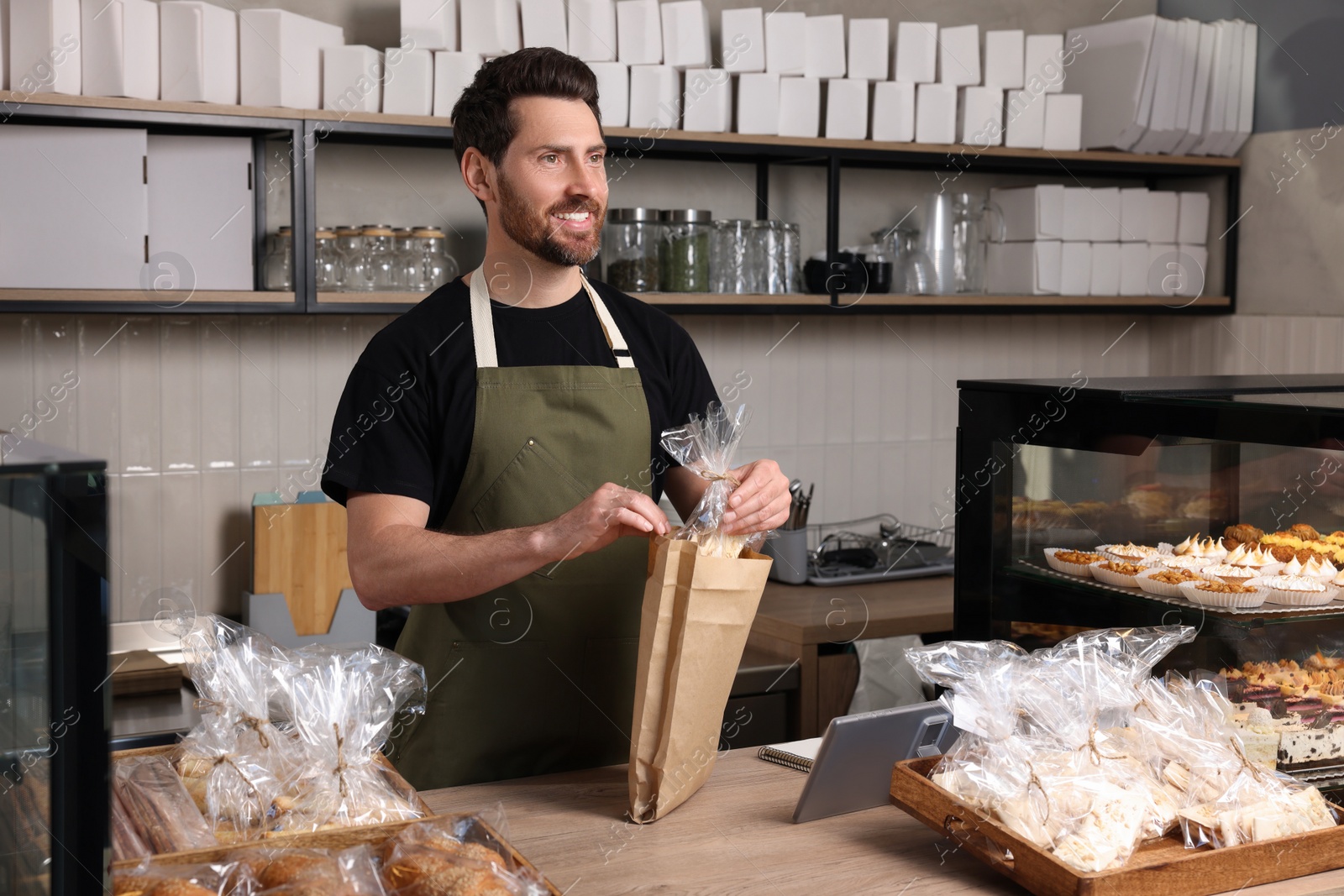 Photo of Happy seller putting pastry into paper bag at cashier desk in bakery shop