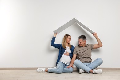 Young family housing concept. Pregnant woman with her husband sitting under cardboard roof on floor indoors. Space for text