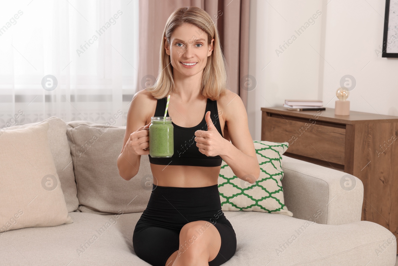 Photo of Young woman in sportswear with mason jar of fresh smoothie at home