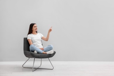 Photo of Beautiful woman pointing at something while sitting in armchair near light grey wall indoors, space for text