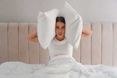 Photo of Unhappy young woman covering ears with pillows in bed at home. Noisy neighbours