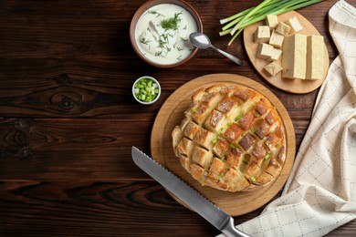 Photo of Freshly baked bread with tofu cheese, green onions, sauce and knife on wooden table, flat lay. Space for text