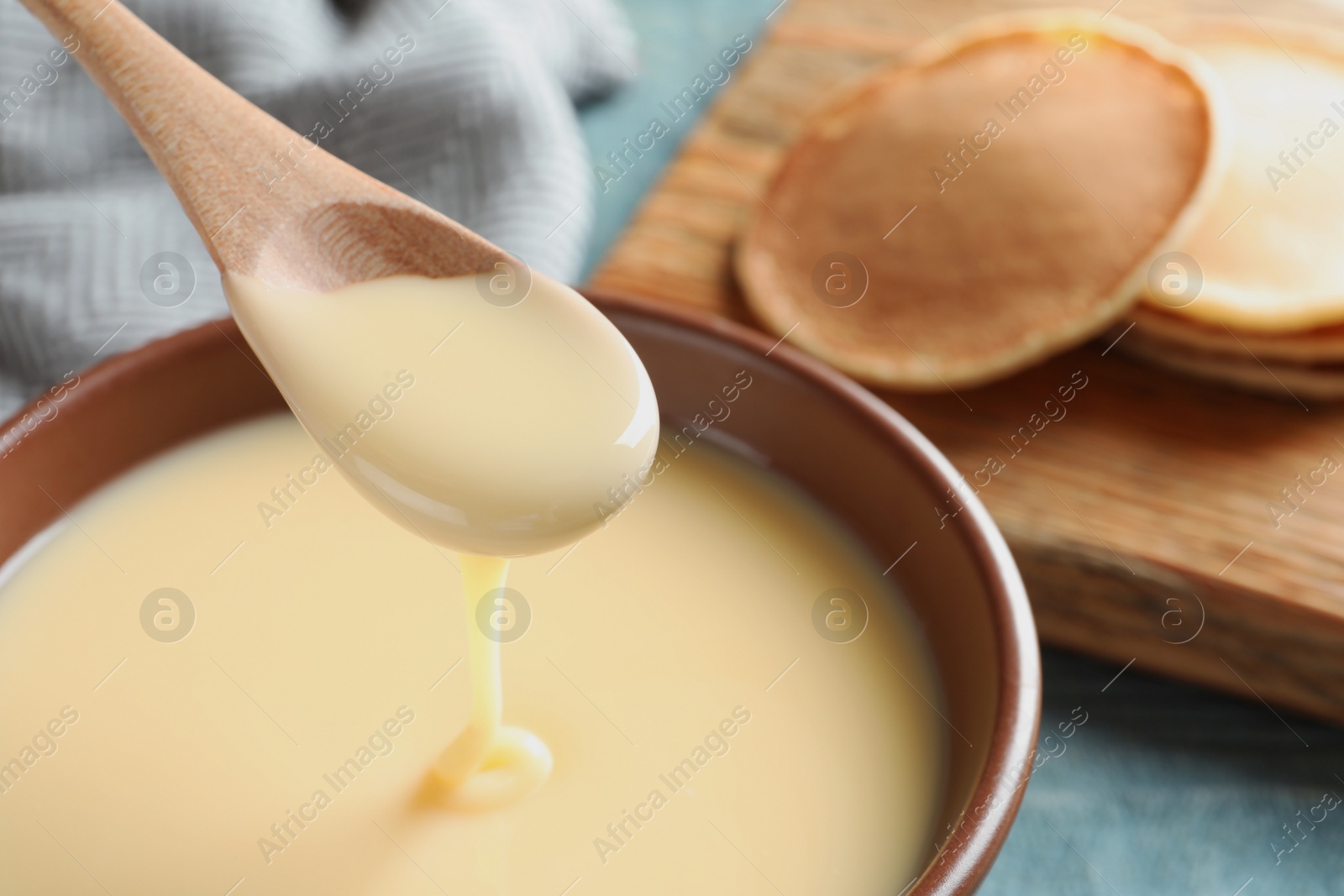 Photo of Spoon of pouring condensed milk over bowl on table, closeup with space for text. Dairy products