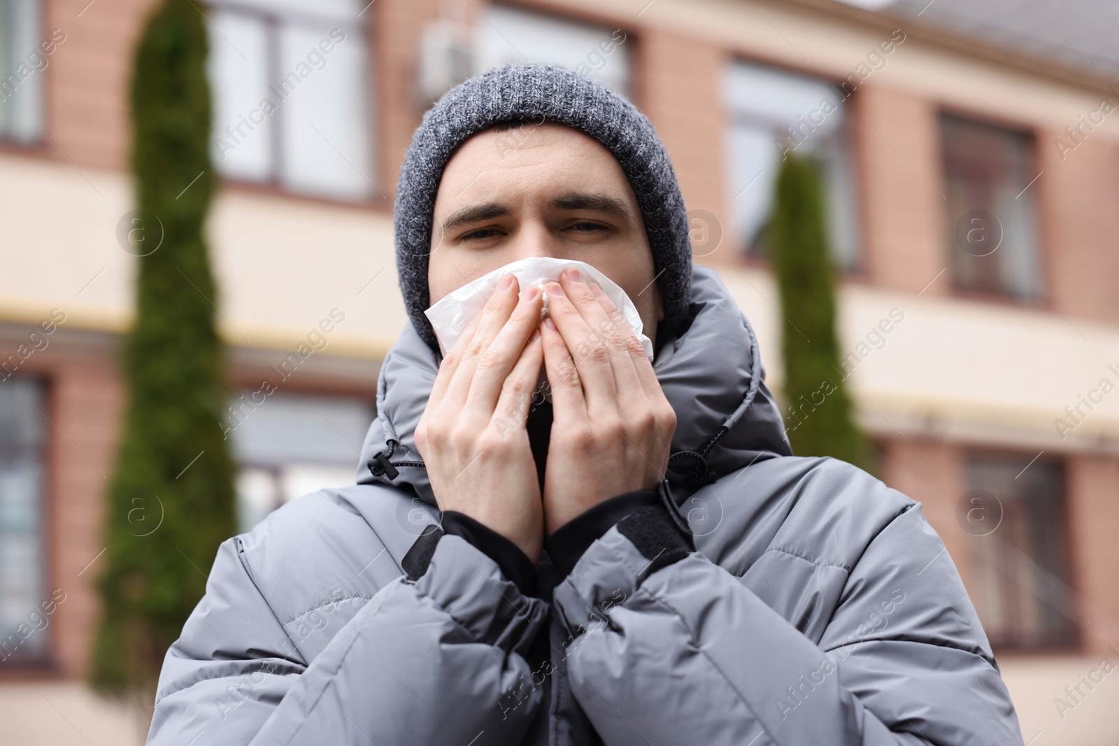 Photo of Sick young man with tissue blowing runny nose outdoors. Cold symptom