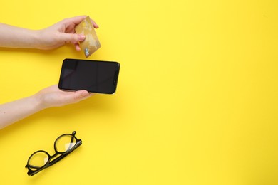 Online payment. Woman with smartphone, credit card and glasses on yellow background, top view. Space for text