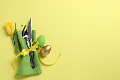 Photo of Cutlery set, painted eggs and beautiful flower on yellow background, flat lay with space for text. Easter celebration