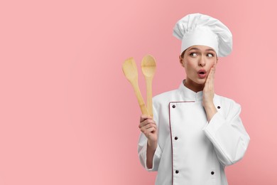 Photo of Surprised chef in uniform holding wooden spatula and spoon on pink background, space for text