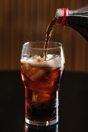 Pouring cola from bottle into glass with ice cubes on table against blurred background