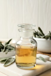 Photo of Bottle of essential sage oil and leaves on white wooden table, closeup