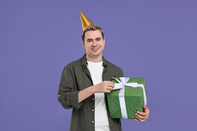 Young man with party hat and gift box on purple background