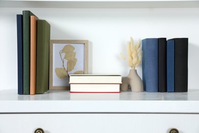 Photo of Hardcover books, picture and vase with dry flowers on white chest of drawers