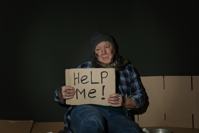 Poor senior man holding cardboard sign HELP ME near dark wall