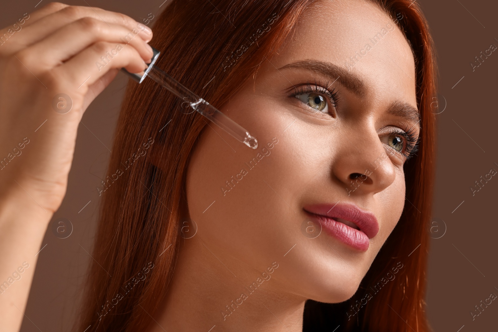 Photo of Beautiful young woman applying cosmetic serum onto her face on brown background, closeup