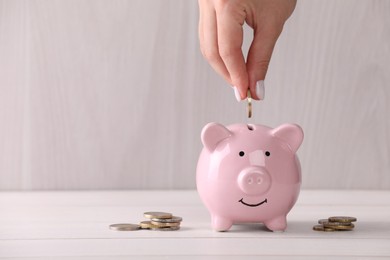 Photo of Woman putting coin into piggy bank at white wooden table, closeup with space for text