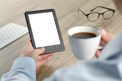 Photo of Man with cup of coffee using e-book reader at wooden table, closeup