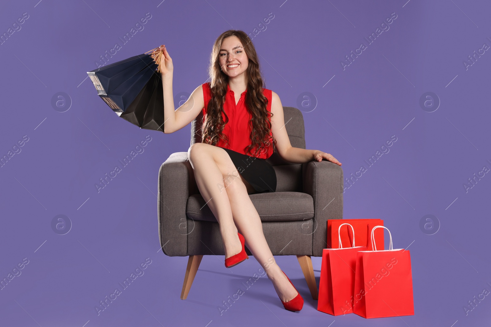 Photo of Happy woman holding paper shopping bags on armchair against purple background