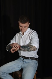 Photo of Young man with tattoos and wristwatch sitting on stool against dark background