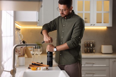 Photo of Romantic dinner. Man opening wine bottle with corkscrew at countertop in kitchen
