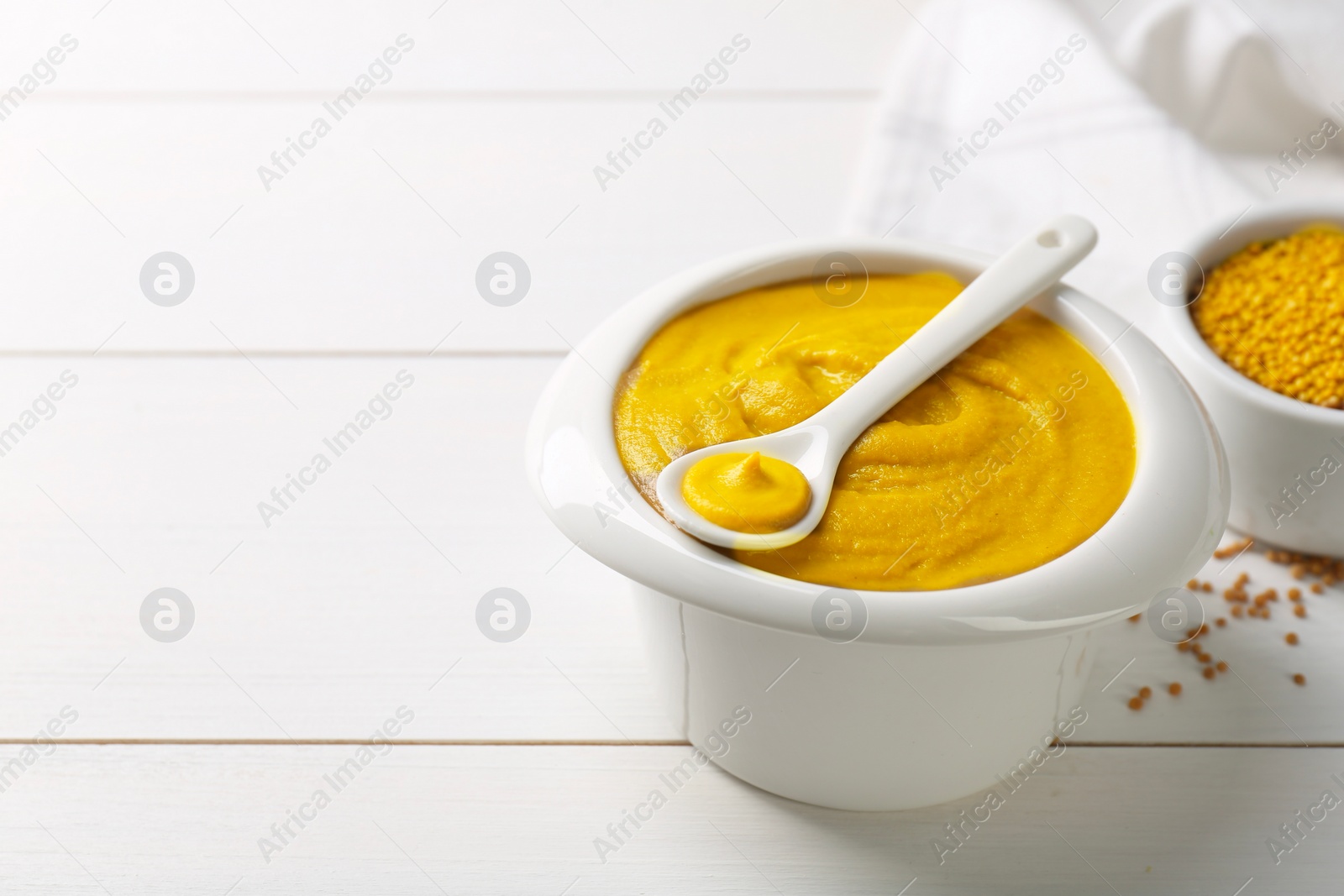 Photo of Spoon and bowl of tasty mustard sauce on white wooden table, space for text