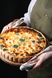 Photo of Woman holding wooden board with delicious homemade salmon quiche on black background, closeup