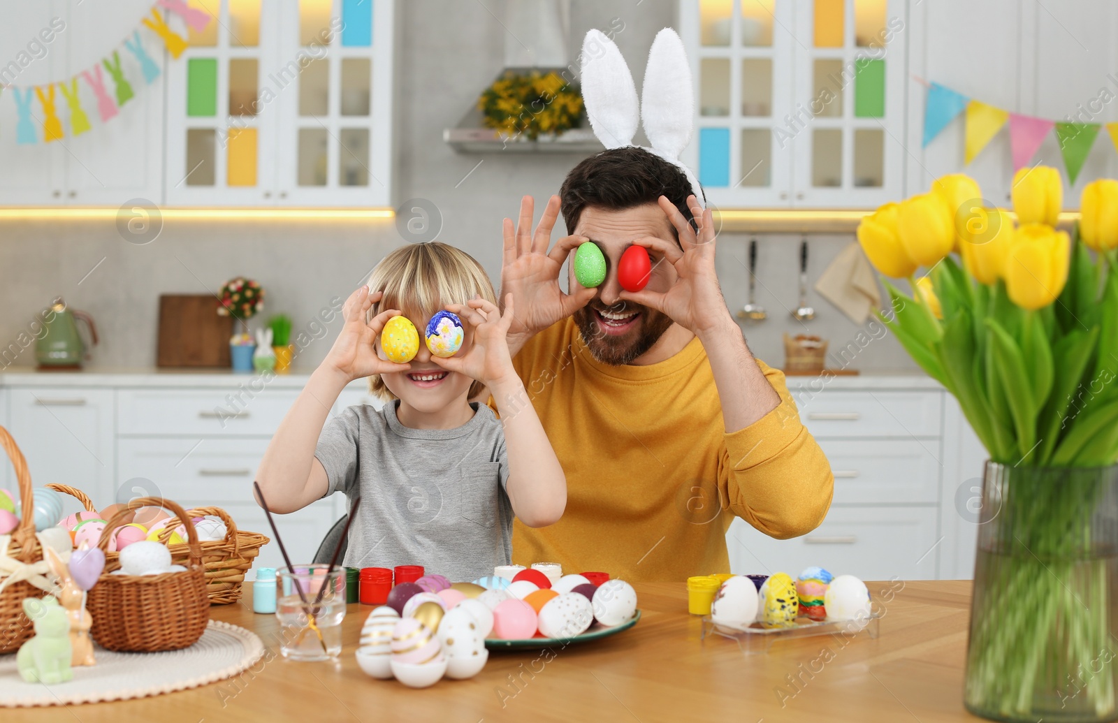 Photo of Father and his cute son covering eyes with Easter eggs at table in kitchen
